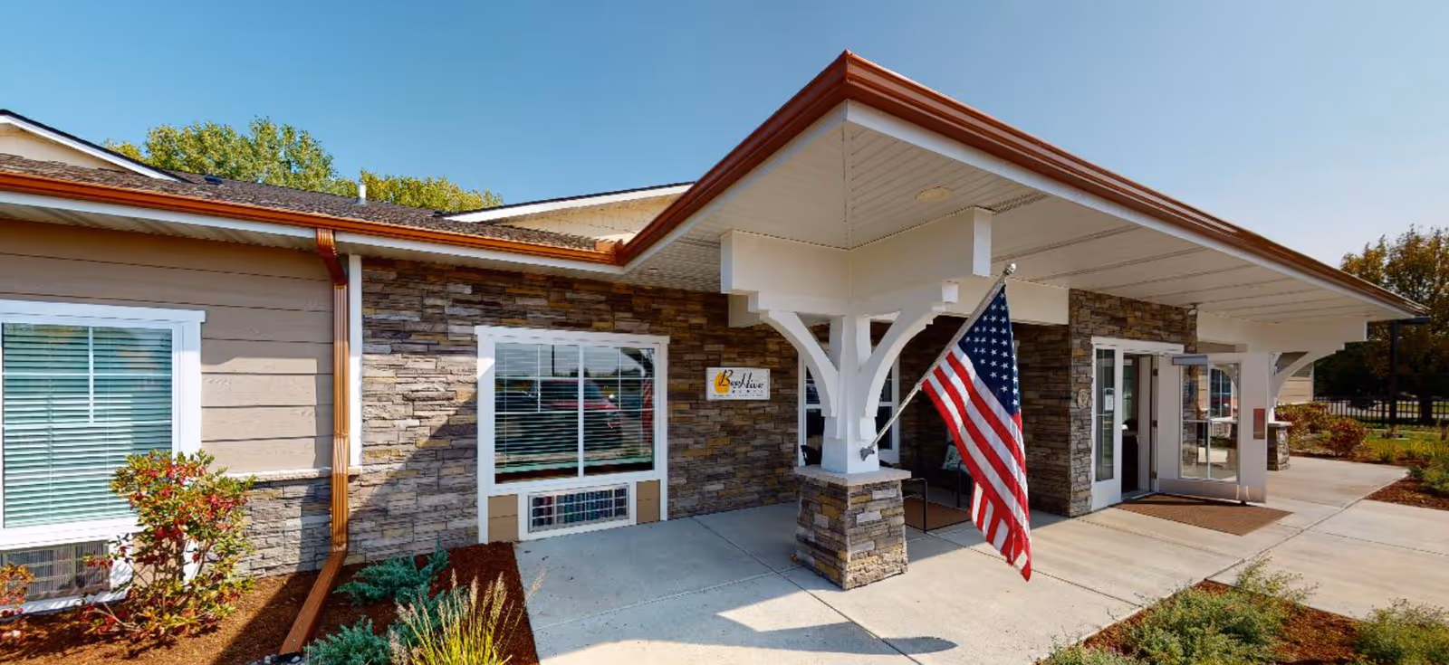 Exterior view of BeeHive Homes of Lakeville South showing the entrance with a covered porch supported by white pillars with stone bases. An American flag is mounted on one of the pillars. The building has stone and beige siding with white trim around the windows and doors. There are plants and landscaping around the entrance area under a clear blue sky.
