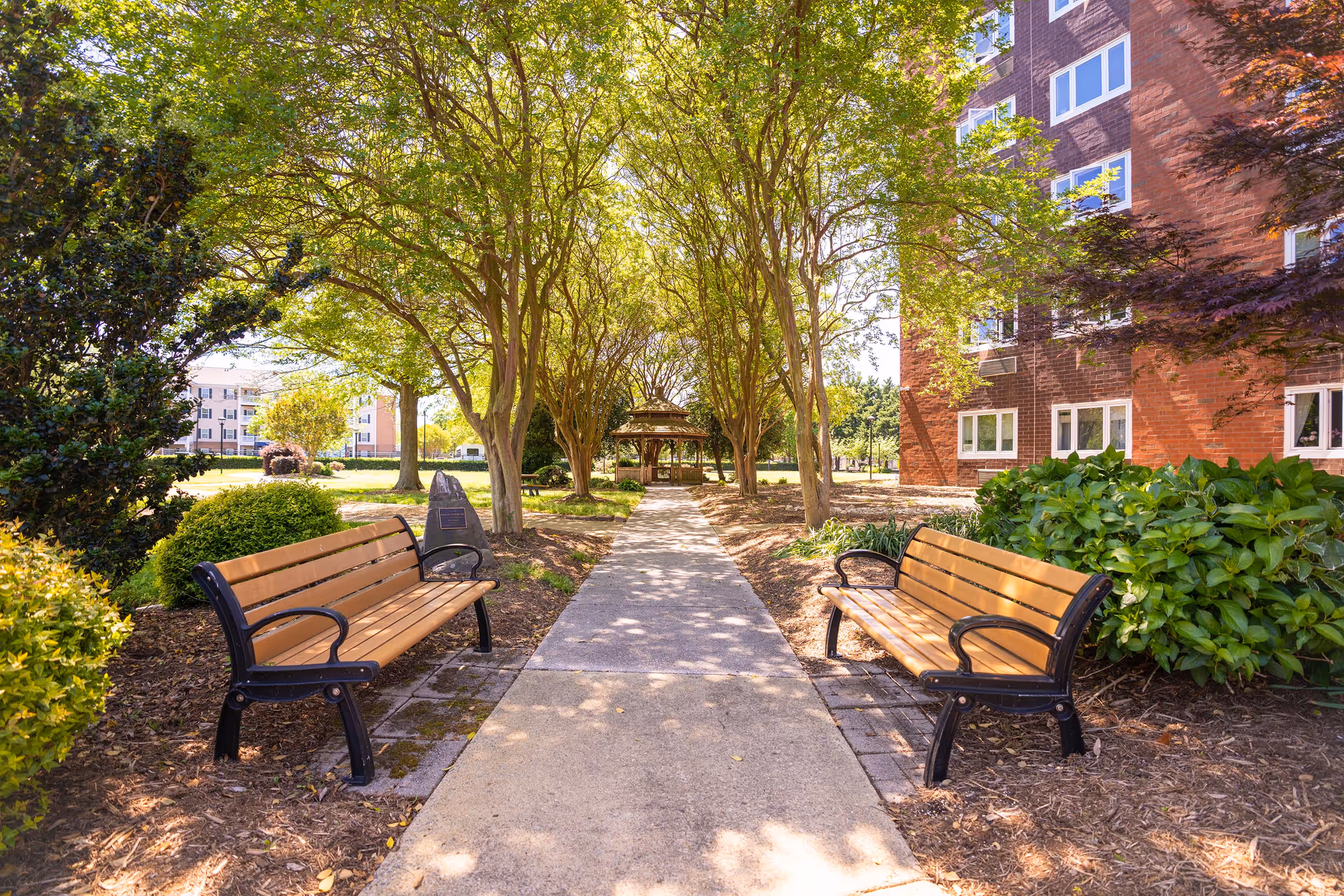 Sunlit outdoor walkway lined with benches and trees leading to a gazebo beside a red-brick building.