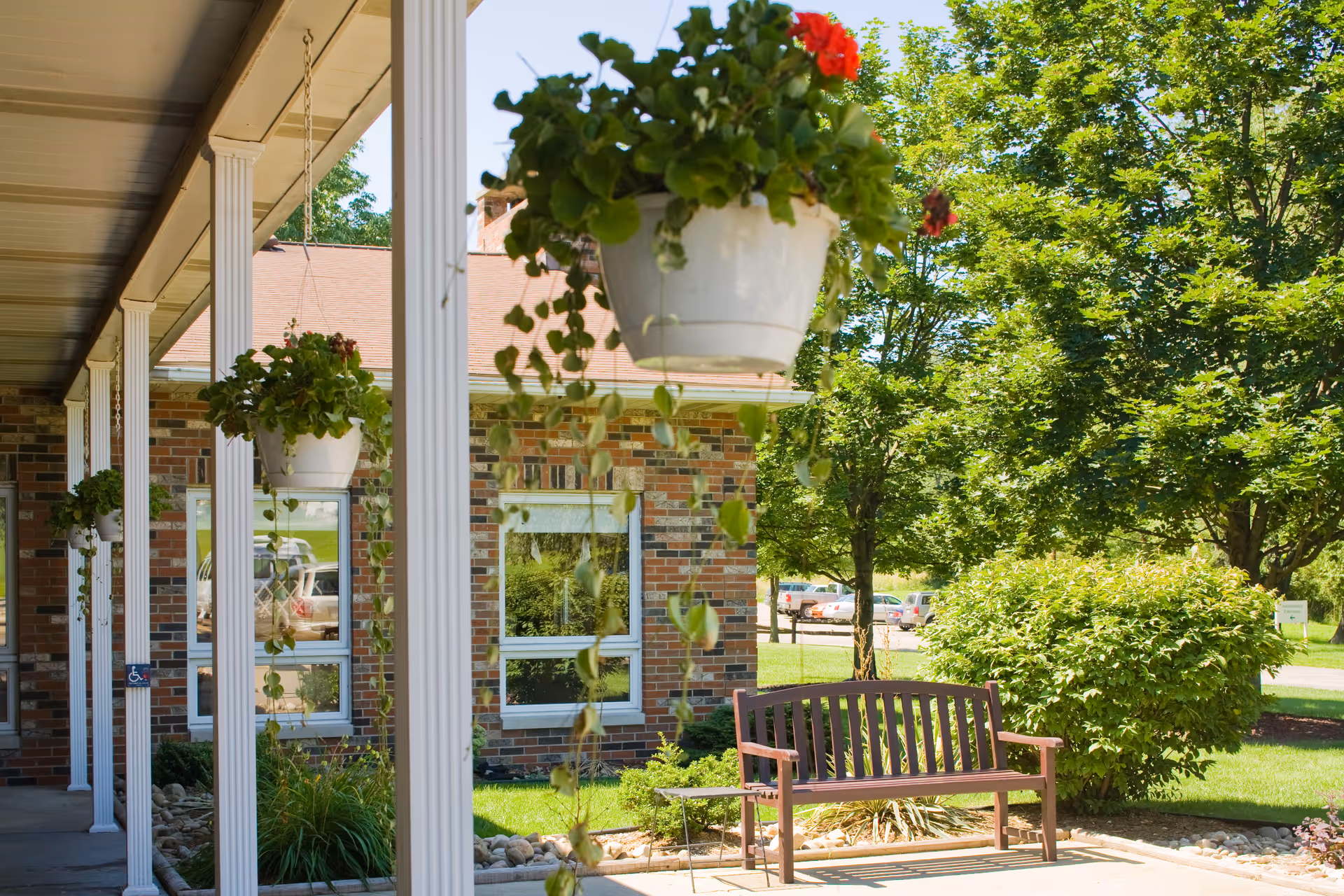 Covered porch with hanging potted plants and a bench outside a brick building surrounded by trees and landscaping.