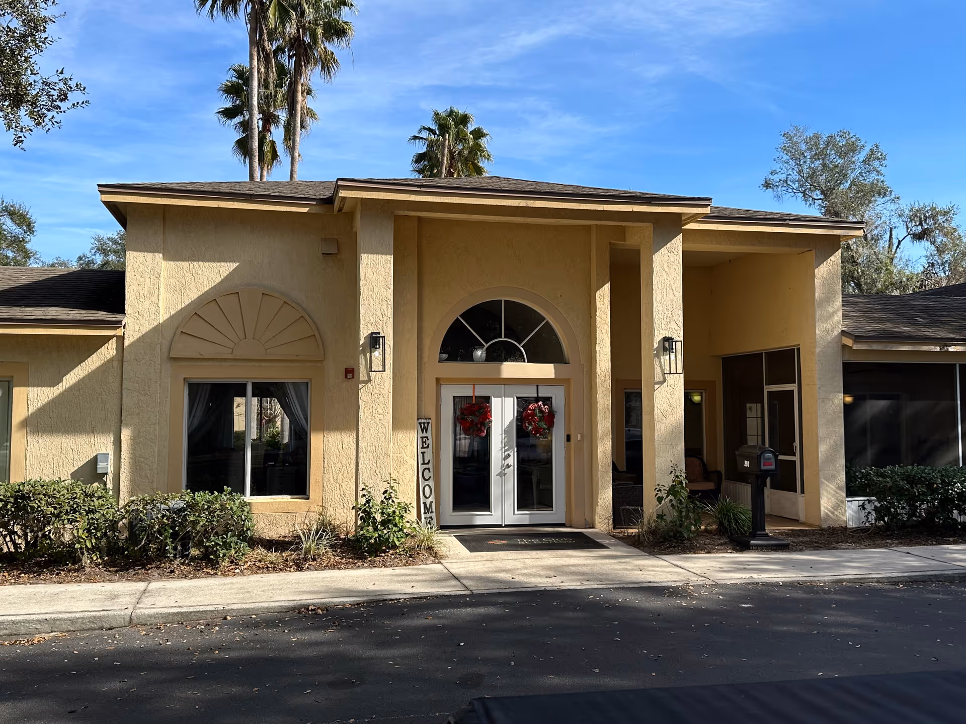 Exterior front entrance of a beige senior living facility building with double glass doors decorated with red wreaths. There is a vertical welcome sign next to the door, some shrubs in front, and tall palm trees in the background under a clear blue sky.
