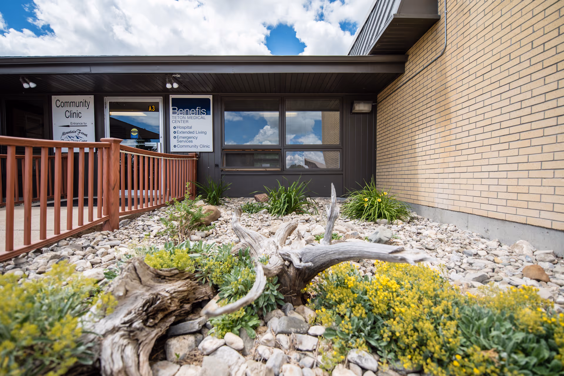 Exterior view of a facility entrance with signs, brown railing, and rocky landscaping.