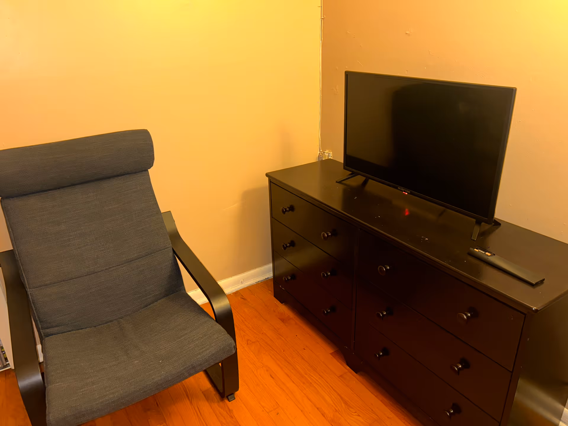 A dark dresser with a flat-screen TV beside a cushioned chair in a small room.