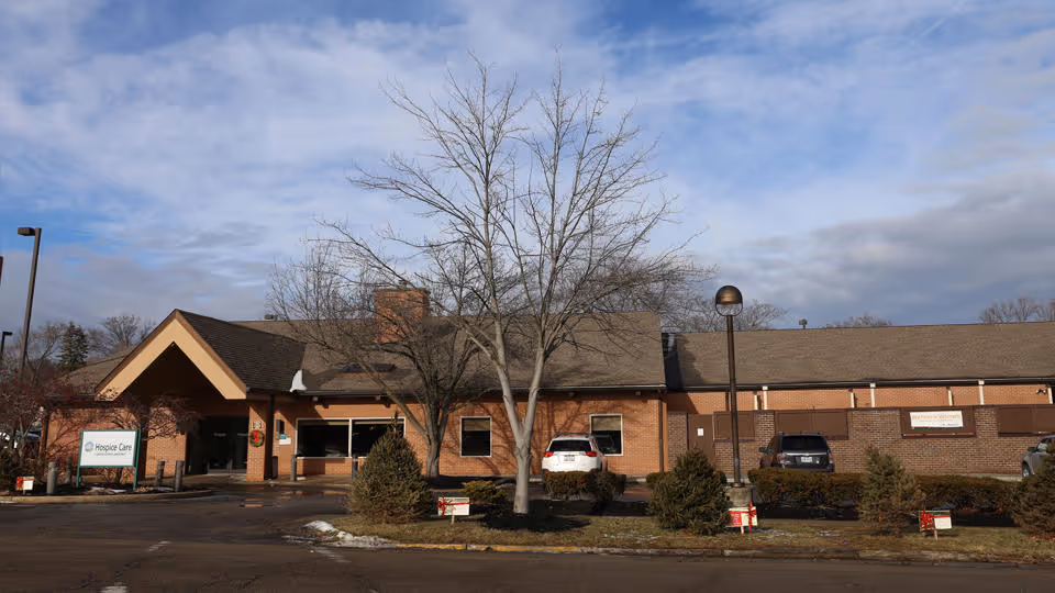 Brick hospice building front with bare trees, parked cars, and an entrance sign near the lot.