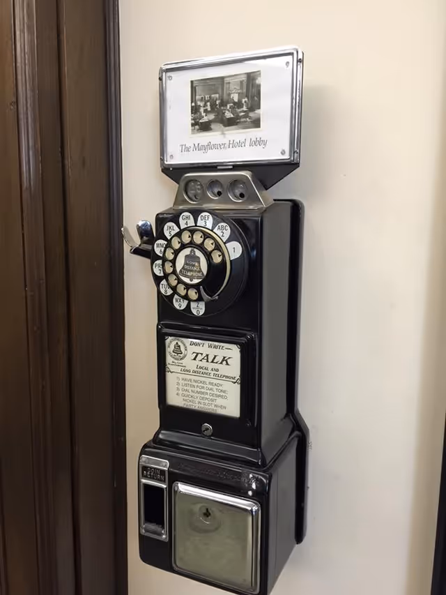 A vintage black rotary payphone mounted on a wall next to a wooden door frame. The phone has a rotary dial with letters and numbers, a coin slot, and instructions for use. Above the phone is a small framed photo labeled 'The Mayflower Hotel lobby.'