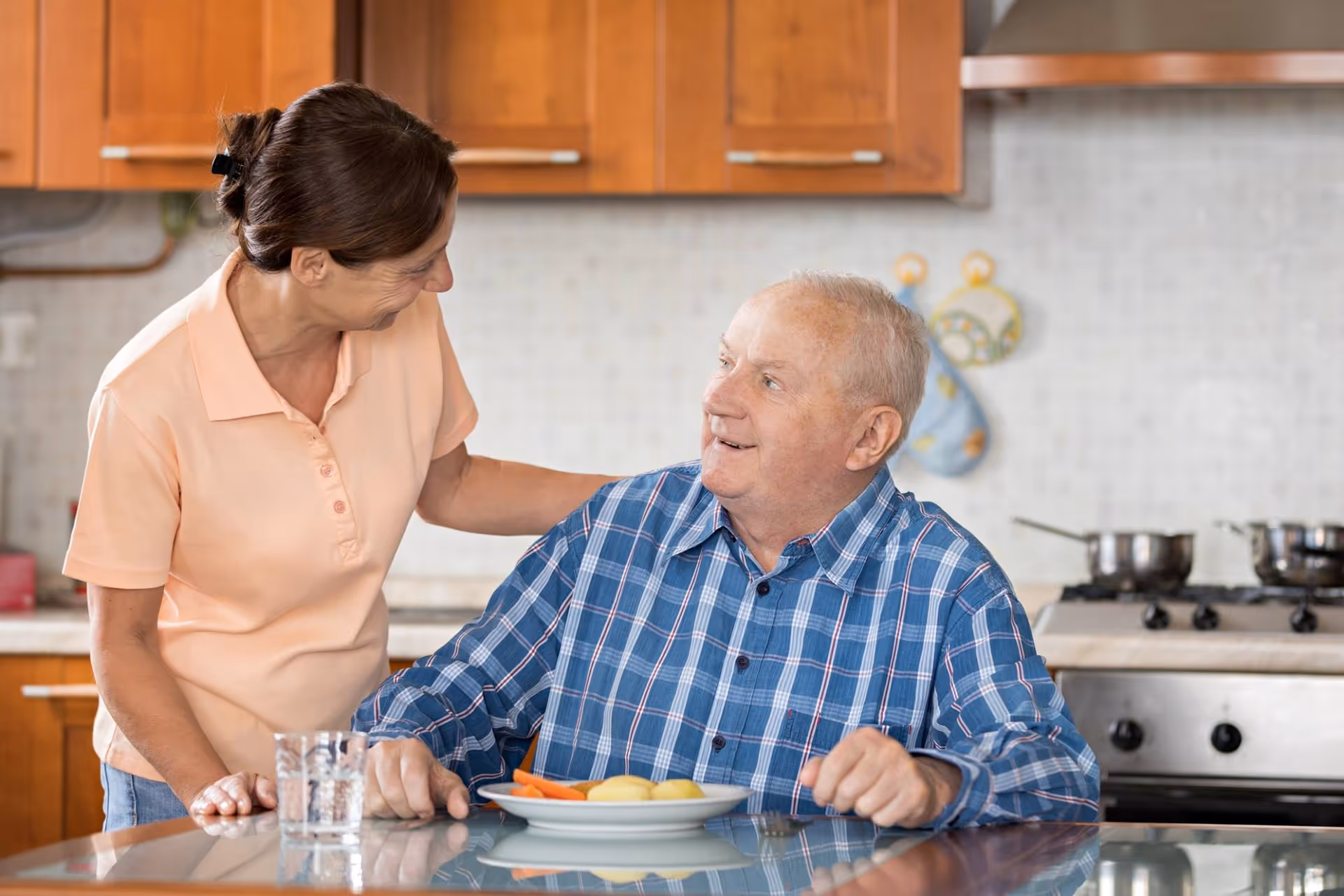 Caregiver sits with elderly man at kitchen counter, sharing a meal together.