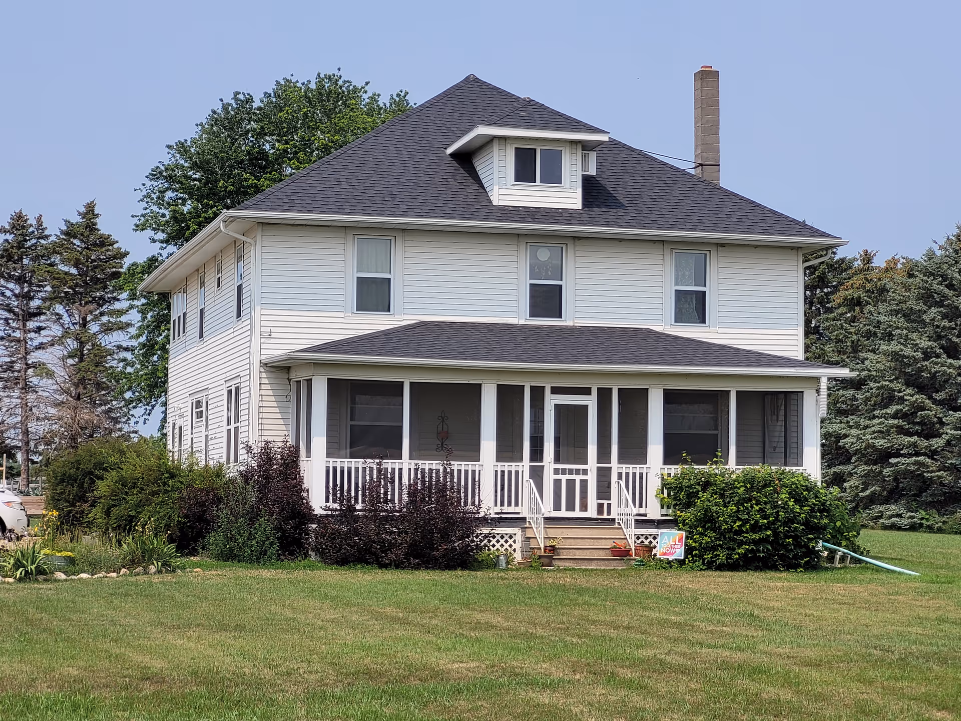 White two-story house with wraparound porch sits on a grassy lawn under blue sky.
