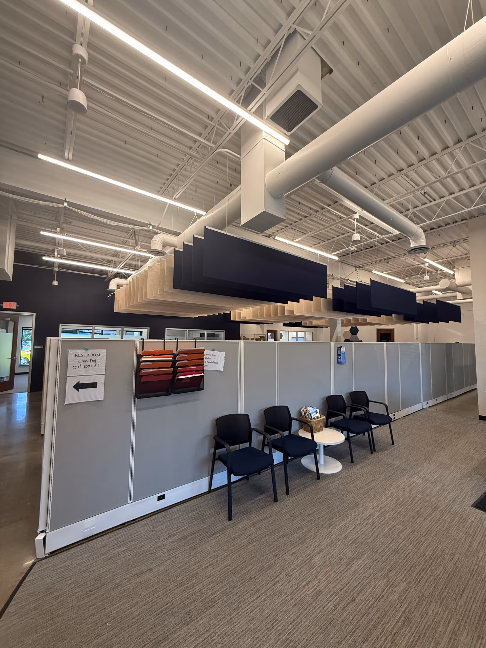 Open interior with gray partitioned cubicles, ceiling panels, and row of chairs.