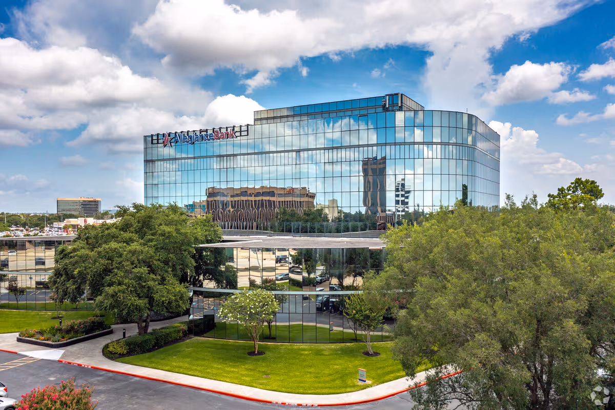 Modern glass office building reflecting the sky, clouds, and surrounding trees.