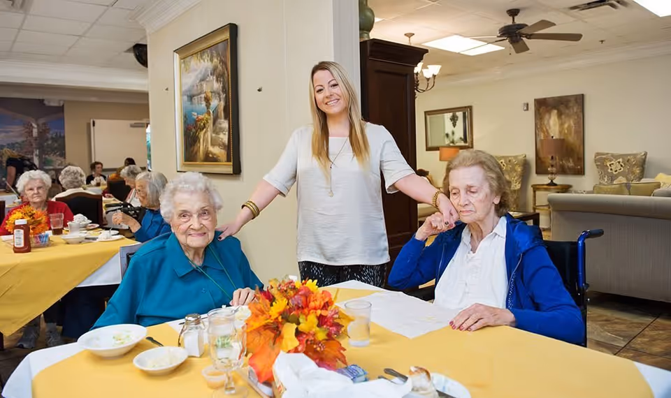 A smiling staff member stands behind two elderly women seated at a decorated dining table in a communal dining room.
