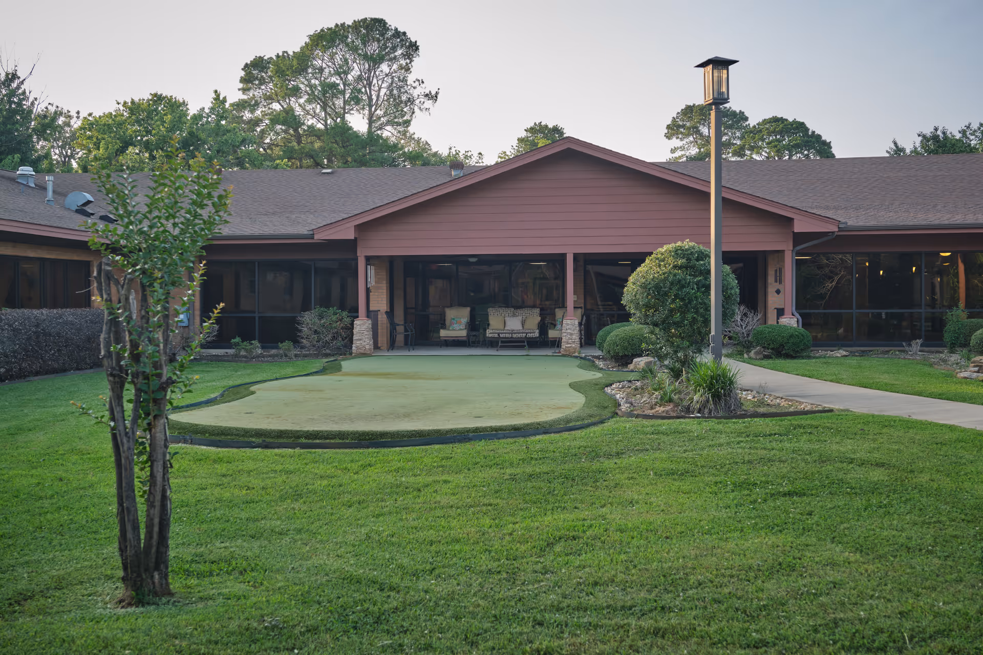 Front exterior of a single-story senior living building with a covered central patio, a small putting green and manicured lawn in the foreground.