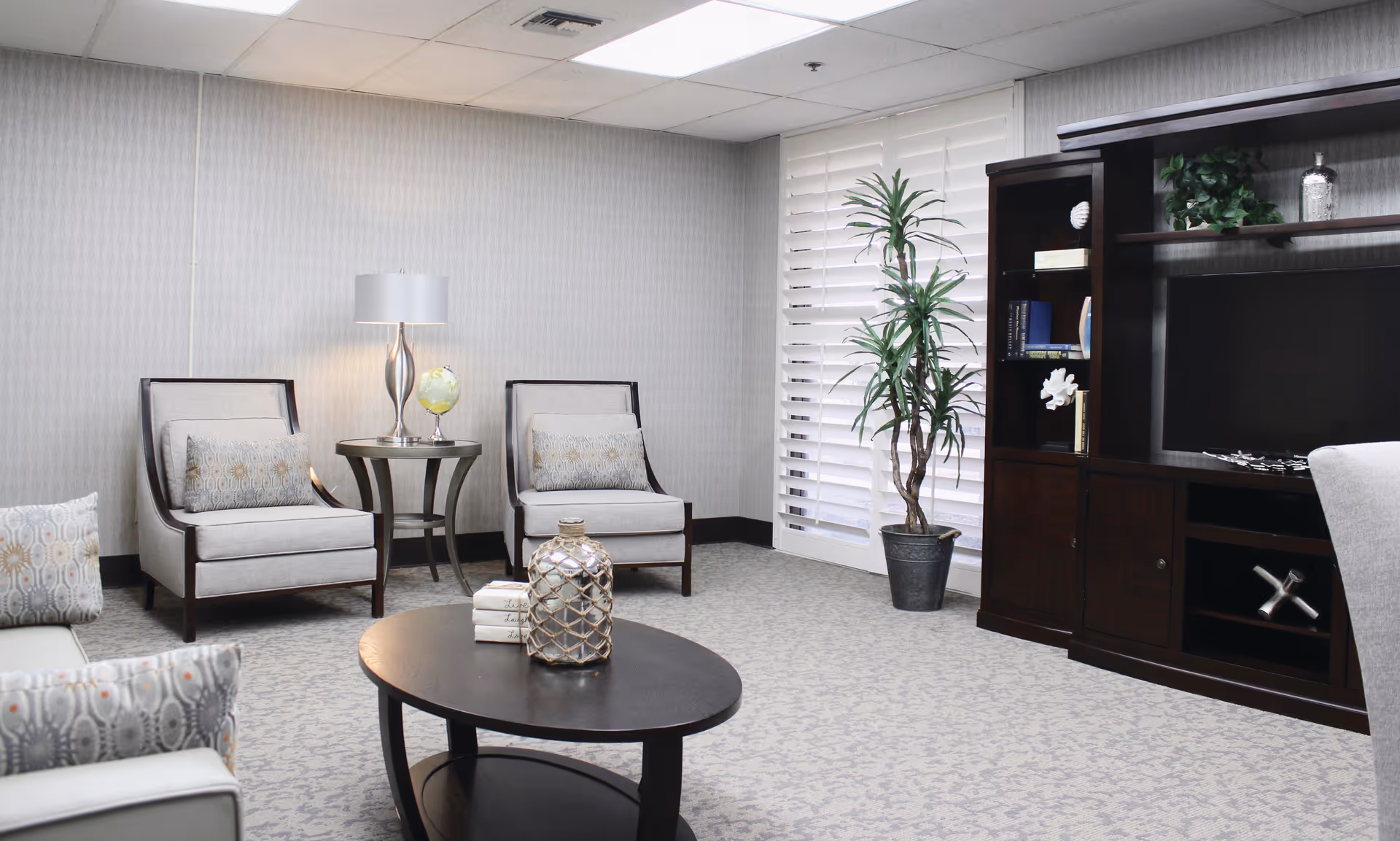 A cozy living room area with two beige armchairs with patterned cushions, a round wooden coffee table with decorative items, a side table with a lamp and a small globe, a tall potted plant near white plantation shutters, and a dark wooden entertainment center with shelves and a TV.