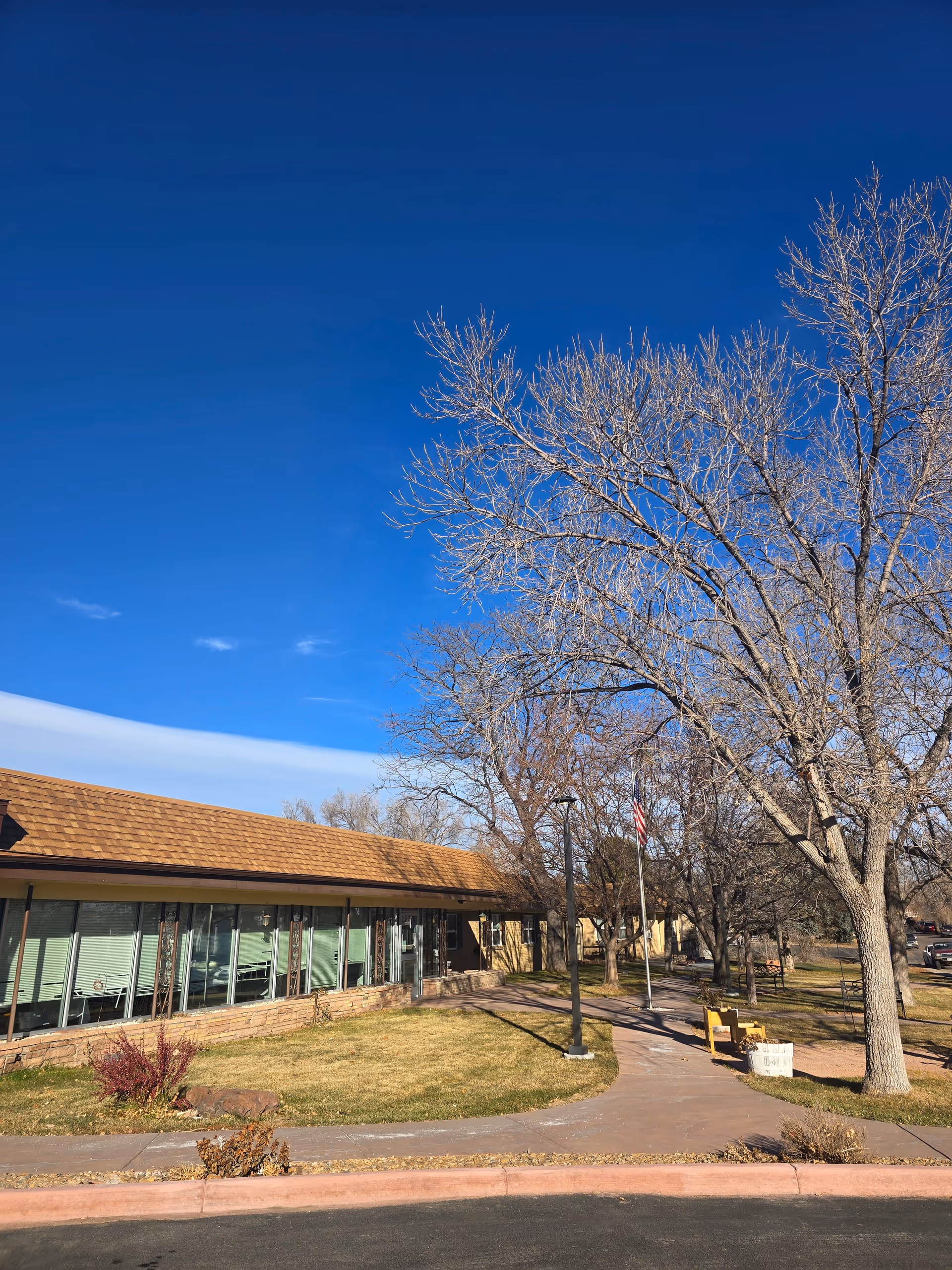 Exterior view of Mapleton Care Center showing a single-story building with large windows, a brown shingled roof, a sidewalk leading to the entrance, leafless trees, a flagpole with an American flag, and a clear blue sky.