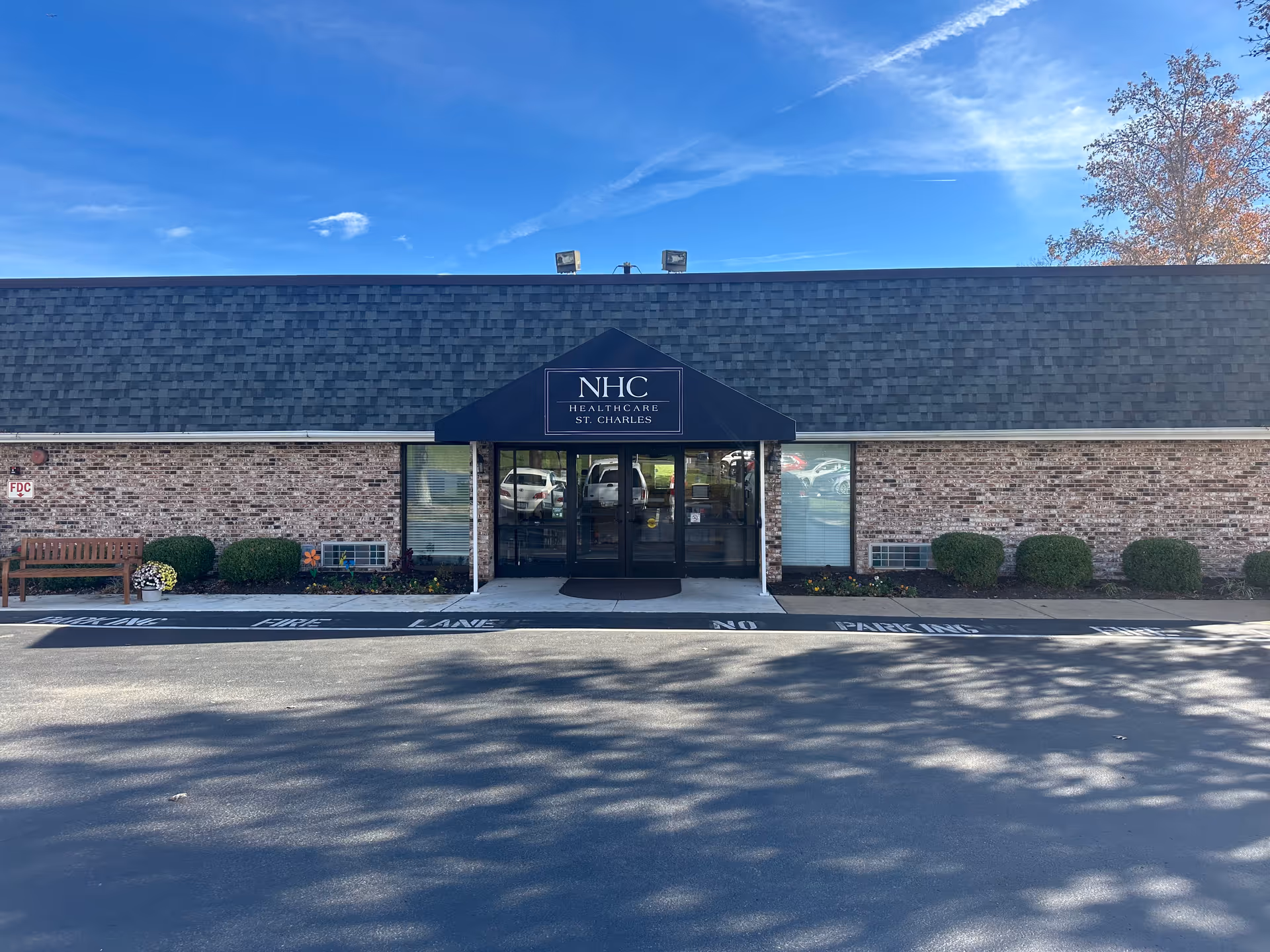 Front entrance of NHC Healthcare - St. Charles, a low brick building with a dark awning and glass double doors.