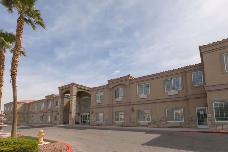 Exterior view of a two-story senior living facility building with beige walls, stone accents, and multiple windows. There are palm trees and a fire hydrant near the driveway leading to the entrance under a partly cloudy sky.