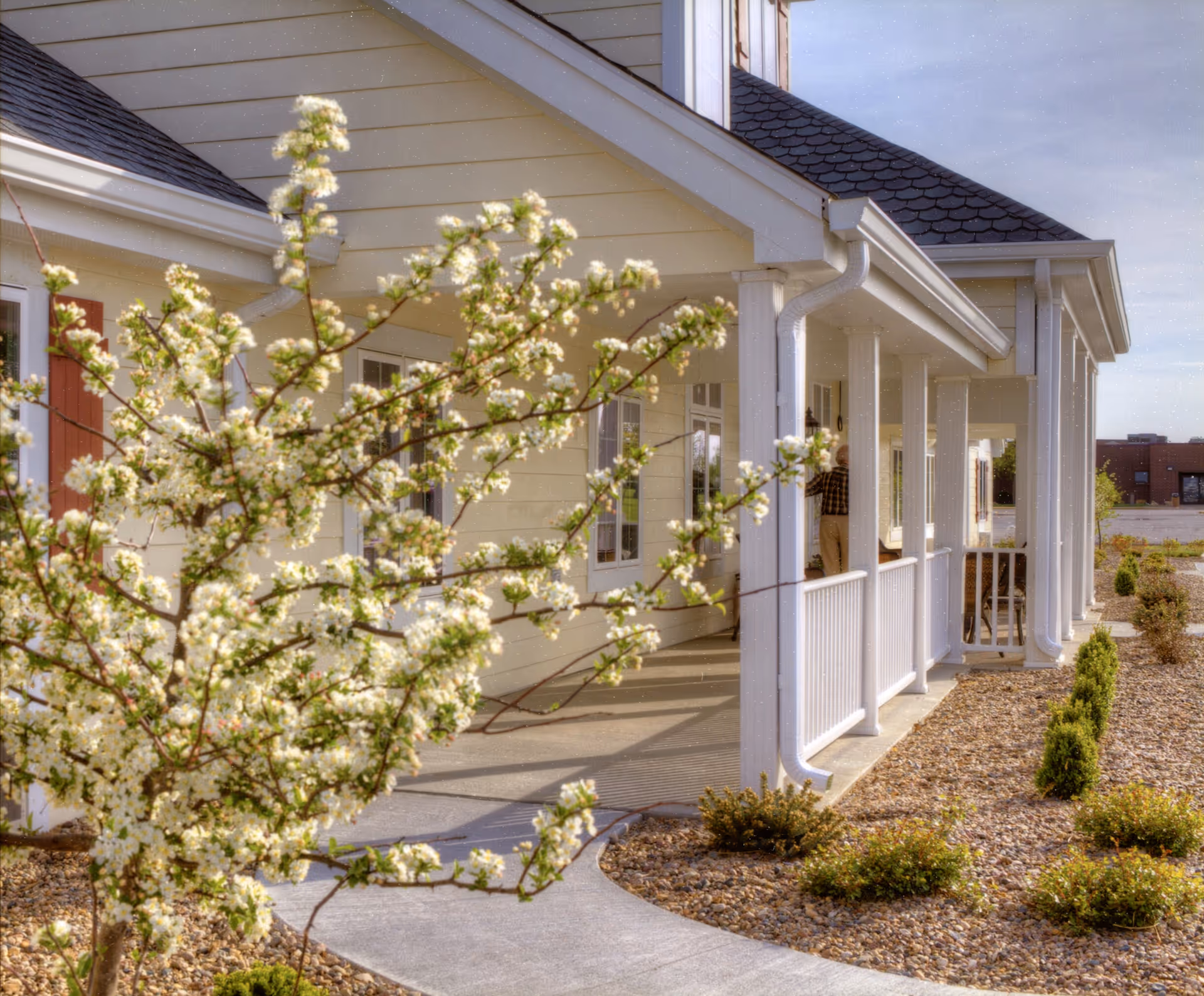 Front porch of a light-colored assisted living building with white columns, a curved walkway, and a blossoming tree in the landscaped front yard.