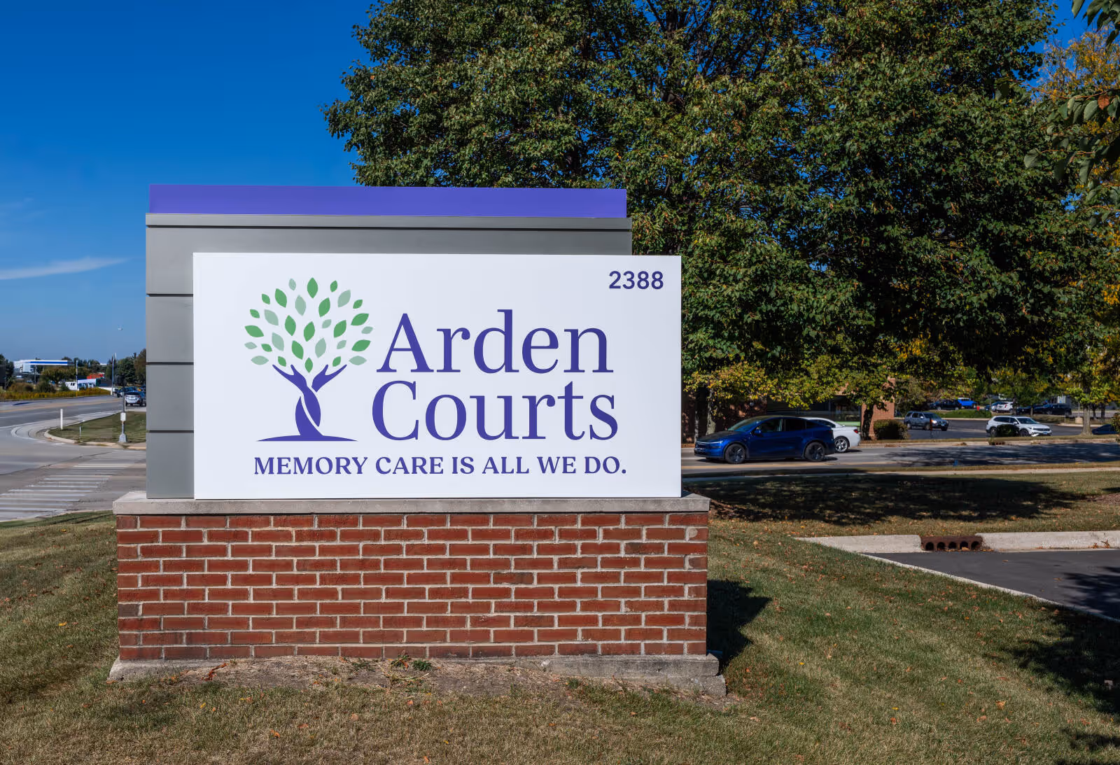 Arden Courts memory care sign on brick pedestal with trees and parking behind.