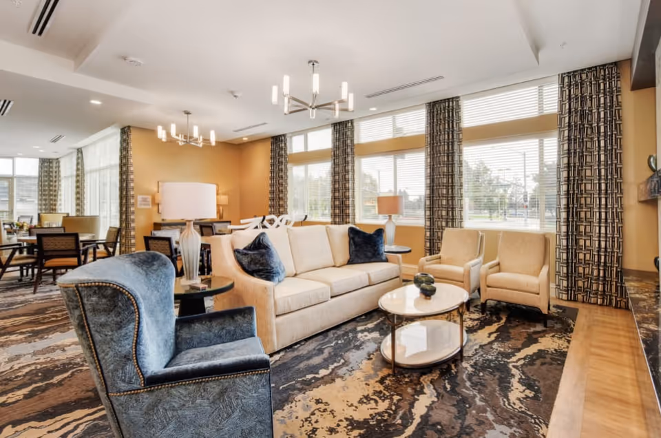 A bright and spacious living room area in a senior living facility with large windows covered by patterned curtains. The room features a beige sofa with dark blue pillows, two beige armchairs, a blue armchair, a round coffee table, and a side table with a lamp. In the background, there are dining tables and chairs, and the room has modern light fixtures on the ceiling.