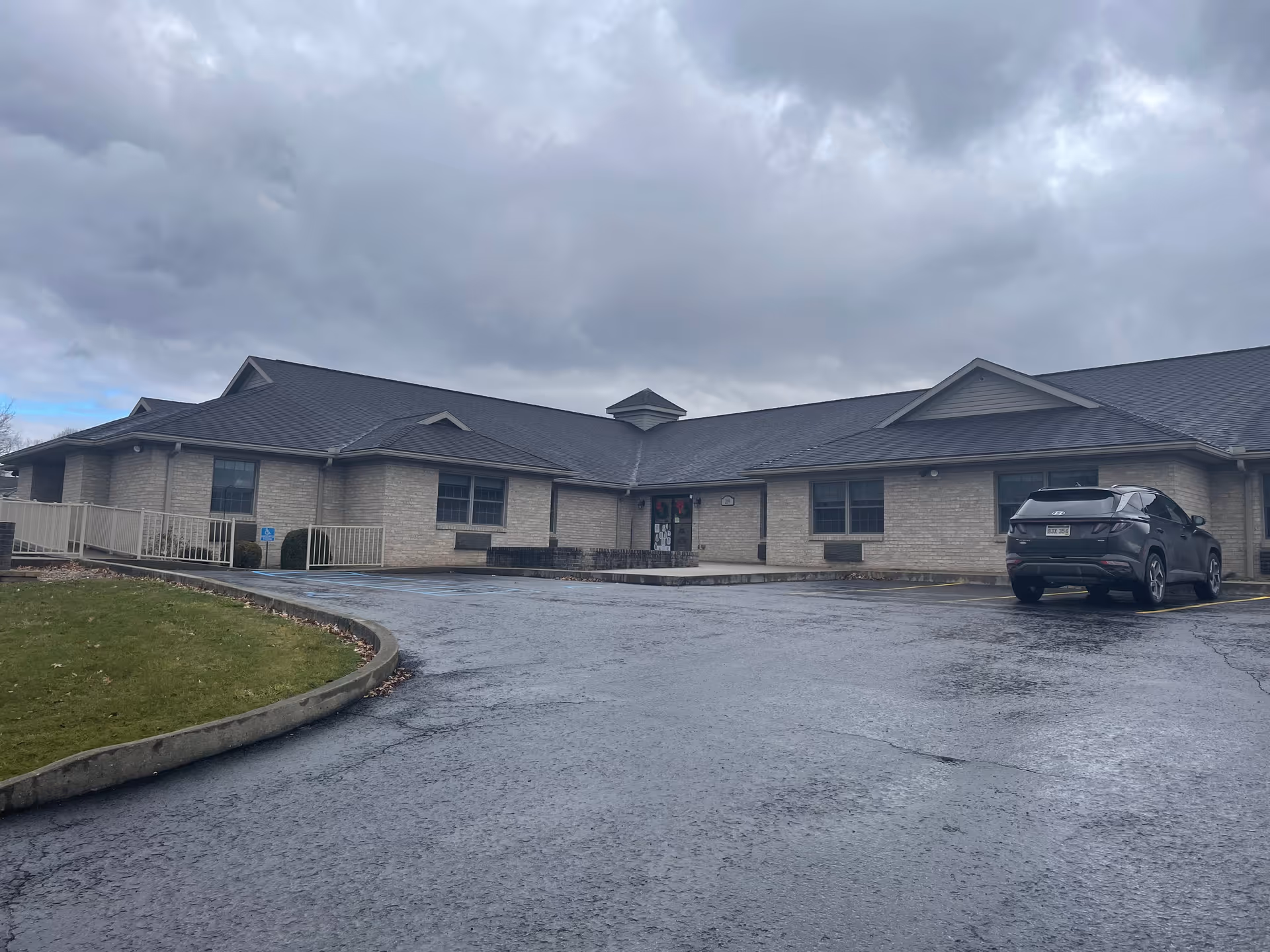 Exterior view of a single-story brick building with a dark shingled roof under a cloudy sky. There is a paved parking lot in front with one parked car and a small grassy area with a curb on the left side. The building has several windows and a main entrance with a small ramp and steps.