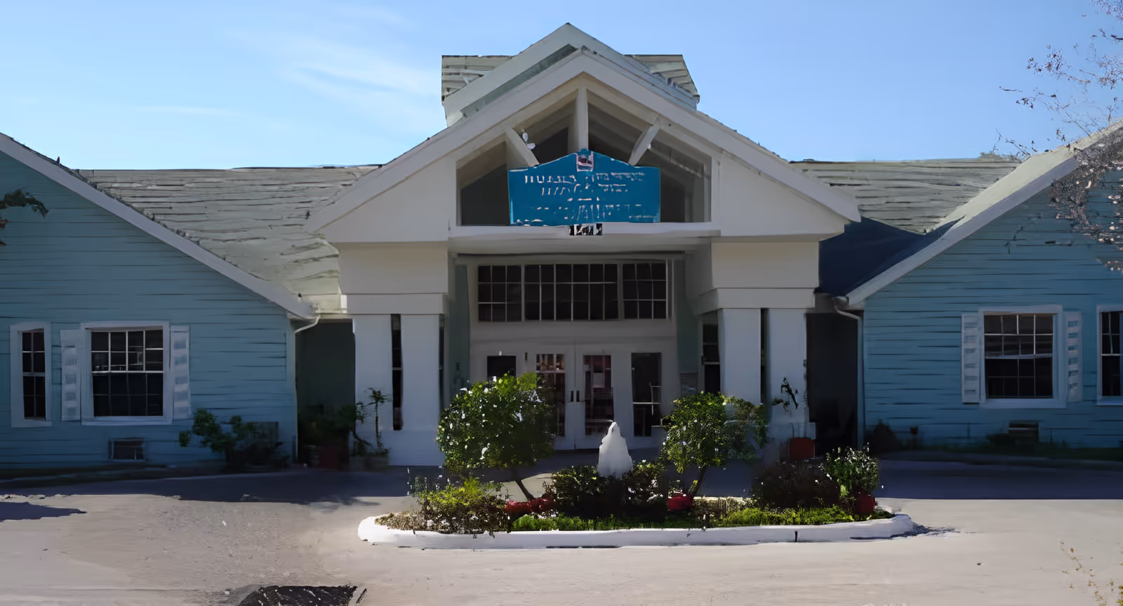 Front exterior view of a single-story building with a peaked roof and a small landscaped area with bushes and a fountain in front. The building has light blue siding with white trim around the windows and entrance.