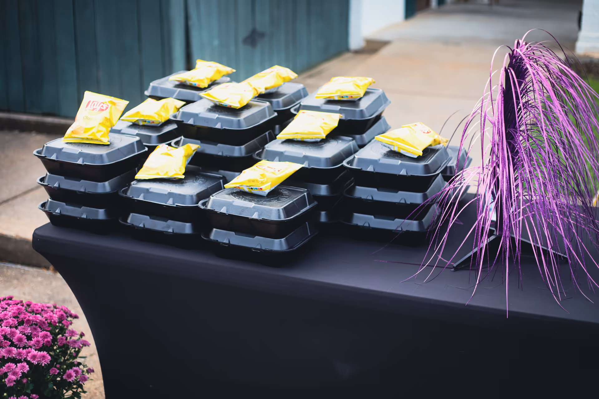 Stacks of packaged meals on a table outside, with purple decorations nearby, ready for distribution.