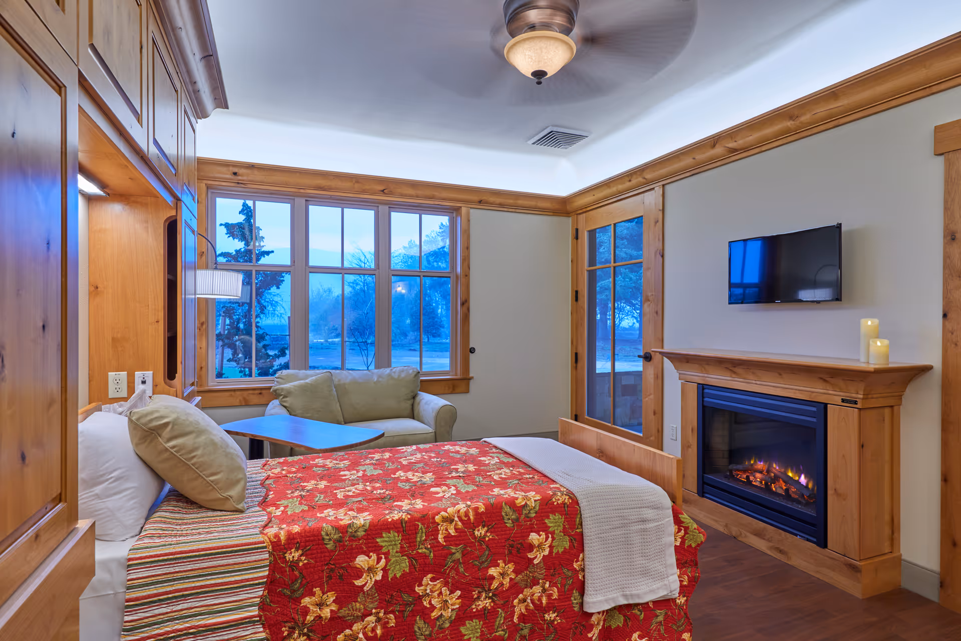 Cozy bedroom suite with a red floral bedspread, sofa, large windows, and a fireplace.