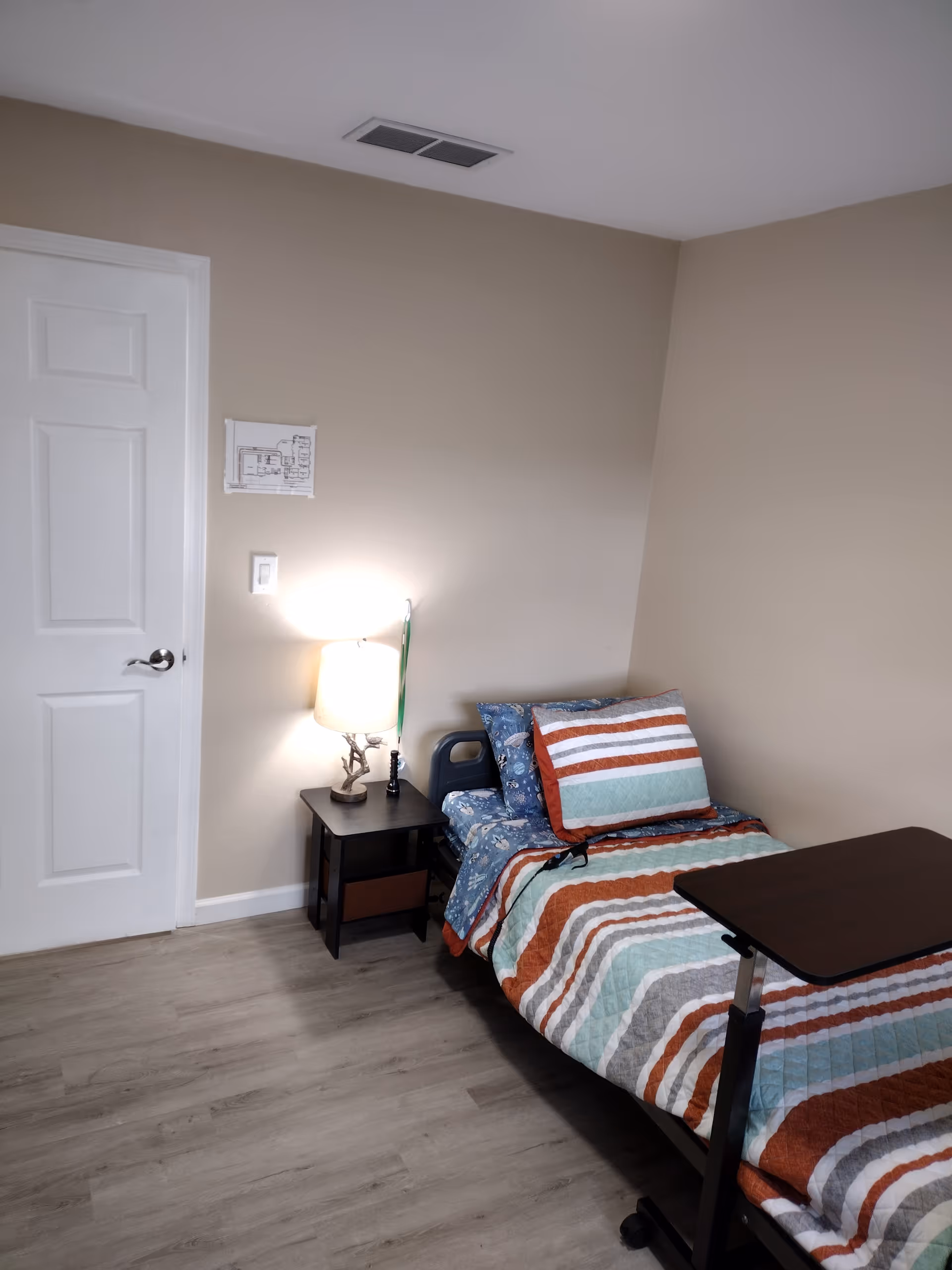 A small furnished bedroom with a single bed, nightstand, lamp, and striped bedding.