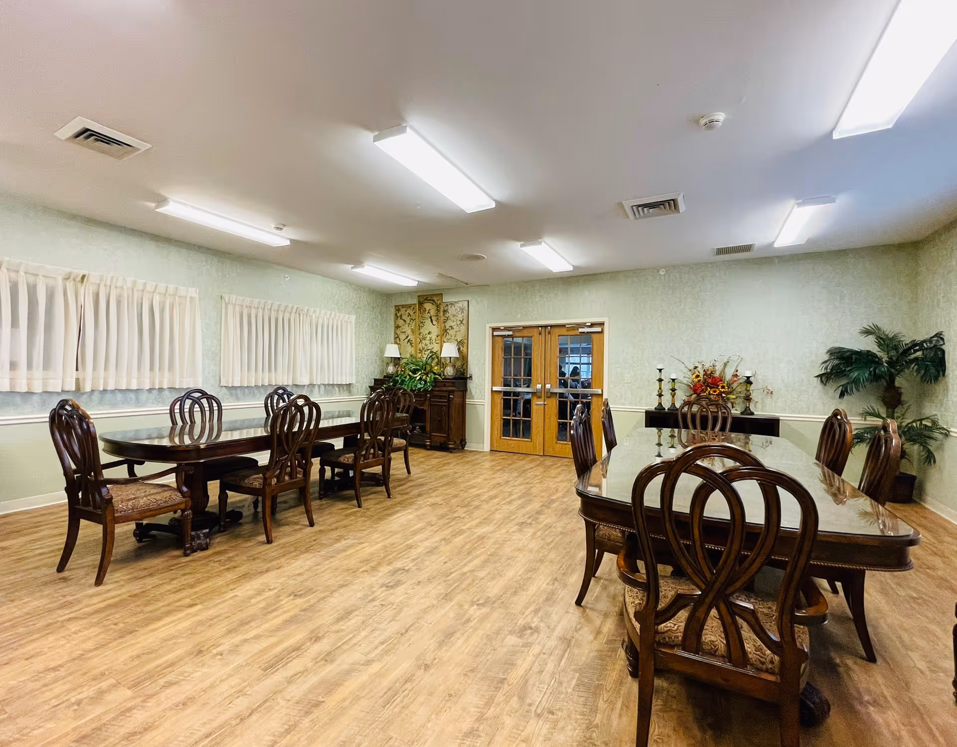 Large dining room with polished wooden tables and chairs, decorative plants, and double doors at the far wall.