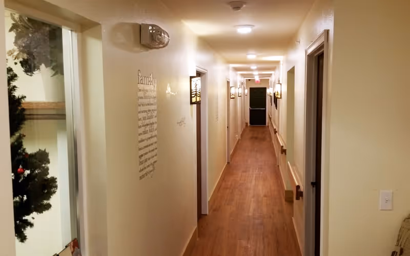 Long, narrow, well-lit interior hallway with wooden floors, wall sconces, handrails and multiple doorways.
