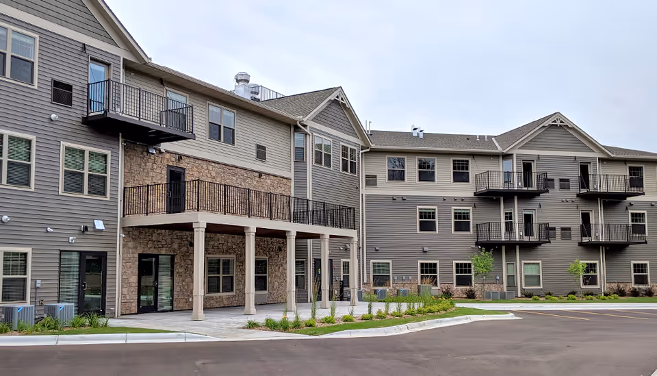 Exterior view of a multi-story senior living facility building with gray siding and stone accents. The building features multiple balconies, windows, and a covered patio area supported by columns. There is a paved driveway and landscaped greenery in front of the building under an overcast sky.