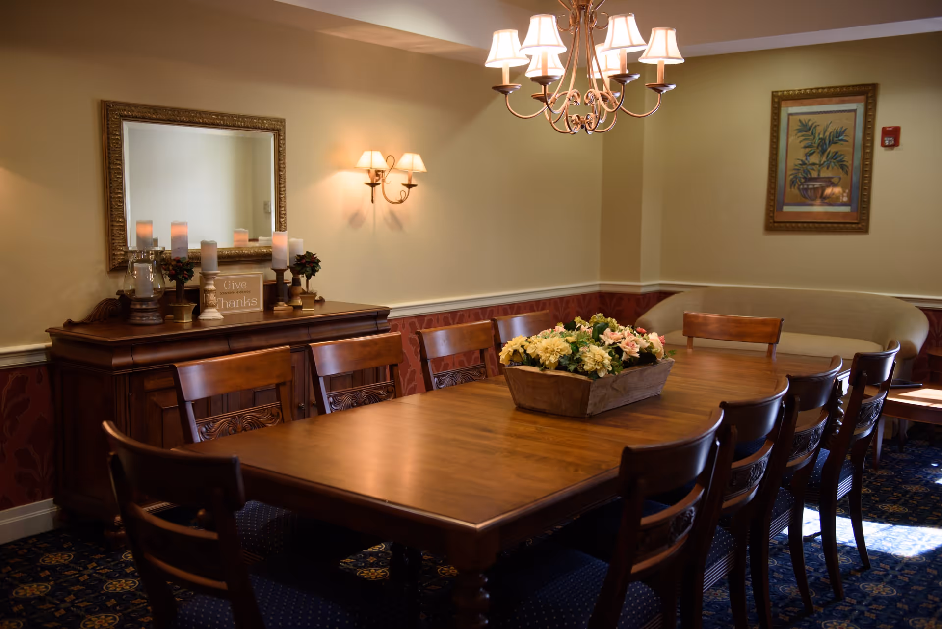 A warmly lit dining room with a large wooden table surrounded by wooden chairs. A floral centerpiece sits on the table. On the left wall, there is a wooden sideboard with candles, small plants, and a sign that reads 'Give thanks' beneath a large framed mirror. A chandelier with multiple lamps hangs above the table, and a framed botanical picture is on the far wall above a beige cushioned bench.