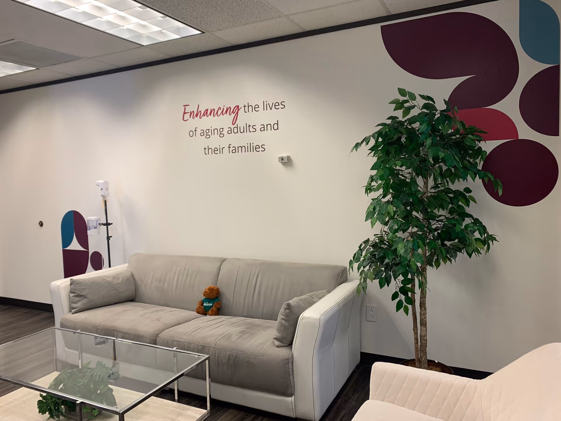 Modern lounge area with gray sofa, glass coffee table, plant, and wall slogan.