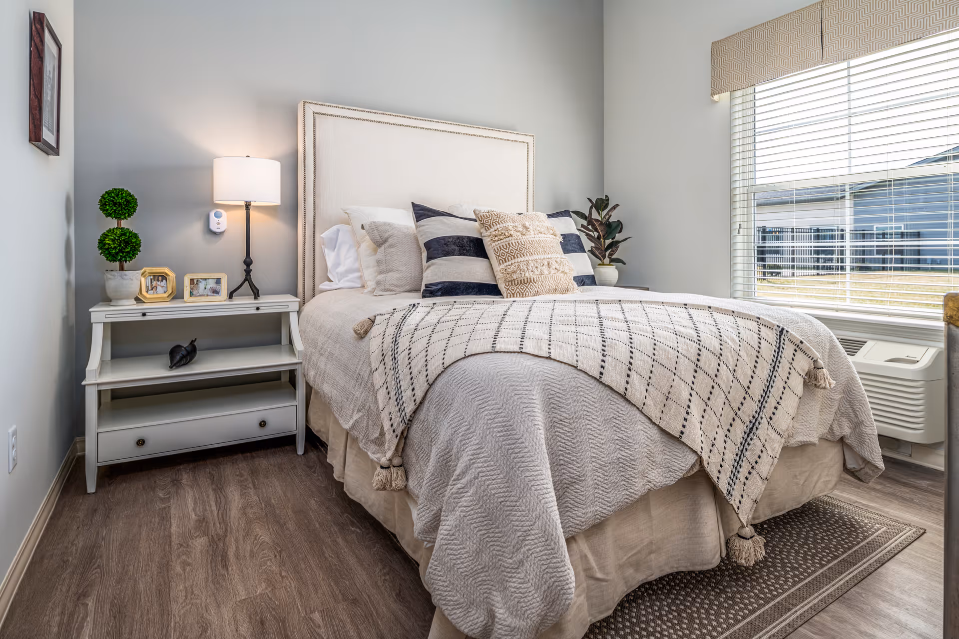 Neatly made bed with patterned throw and pillows beside a white nightstand and lamp in a bright, neutral-toned bedroom with a window.