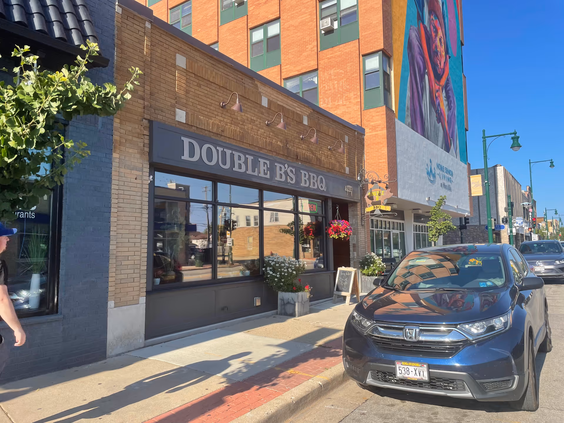 Sunny street storefronts line a downtown block, featuring a barbecue restaurant and a parked car.