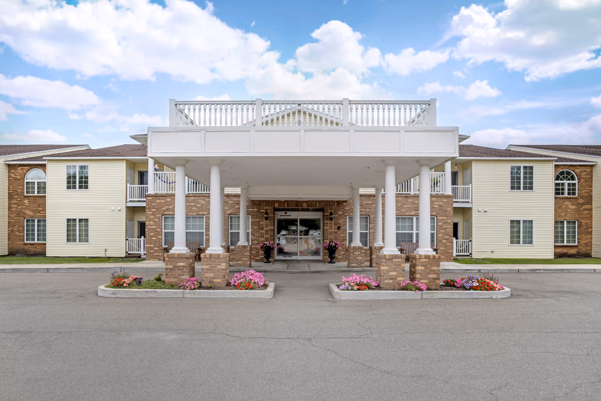Front exterior view of Brookdale Castle Gardens facility showing a covered entrance with white columns and brick bases, flower beds on either side, and a two-story building with windows and balconies under a partly cloudy sky.