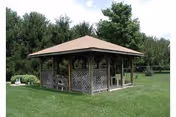 A wooden gazebo with lattice sides sits on a grassy lawn among trees.
