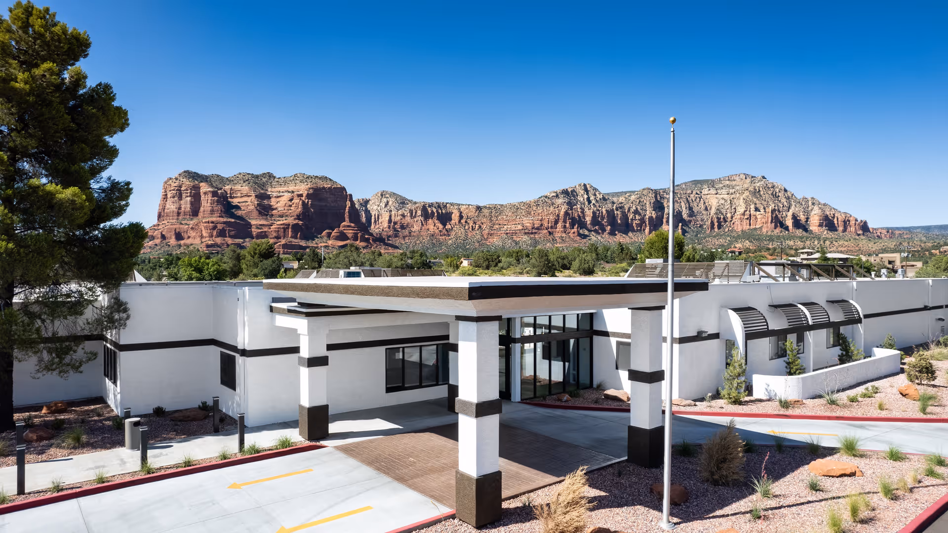 Exterior view of a single-story white building with black trim, featuring a covered entrance and large windows. The building is surrounded by desert landscaping with rocks and sparse vegetation. In the background, there are large red rock formations under a clear blue sky.