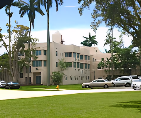 Exterior view of a multi-story beige building surrounded by green grass, tall trees, and several parked cars under a clear blue sky.