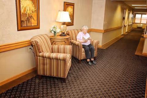 An elderly resident sits reading a magazine in a striped armchair along the Sunrise Villa hallway.