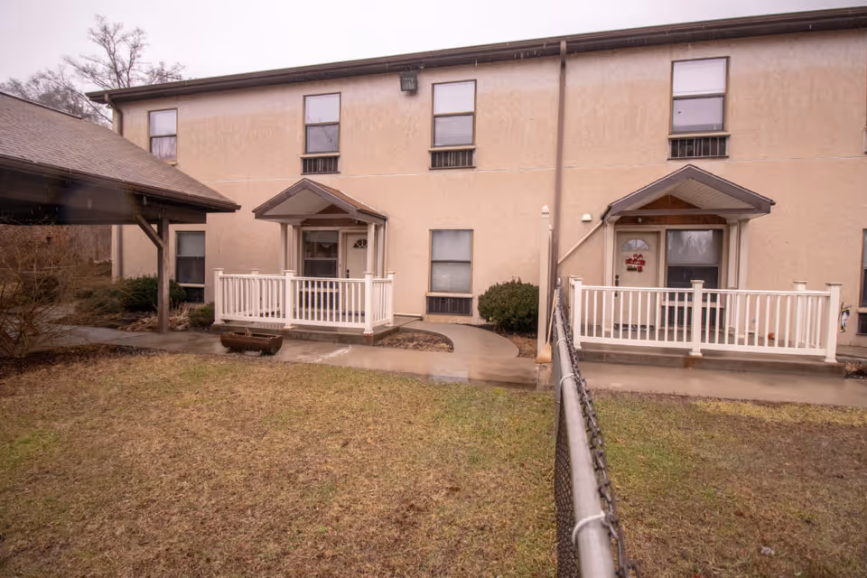 Two-story beige residential building with two covered front porches, white railings, and a fenced grassy yard.