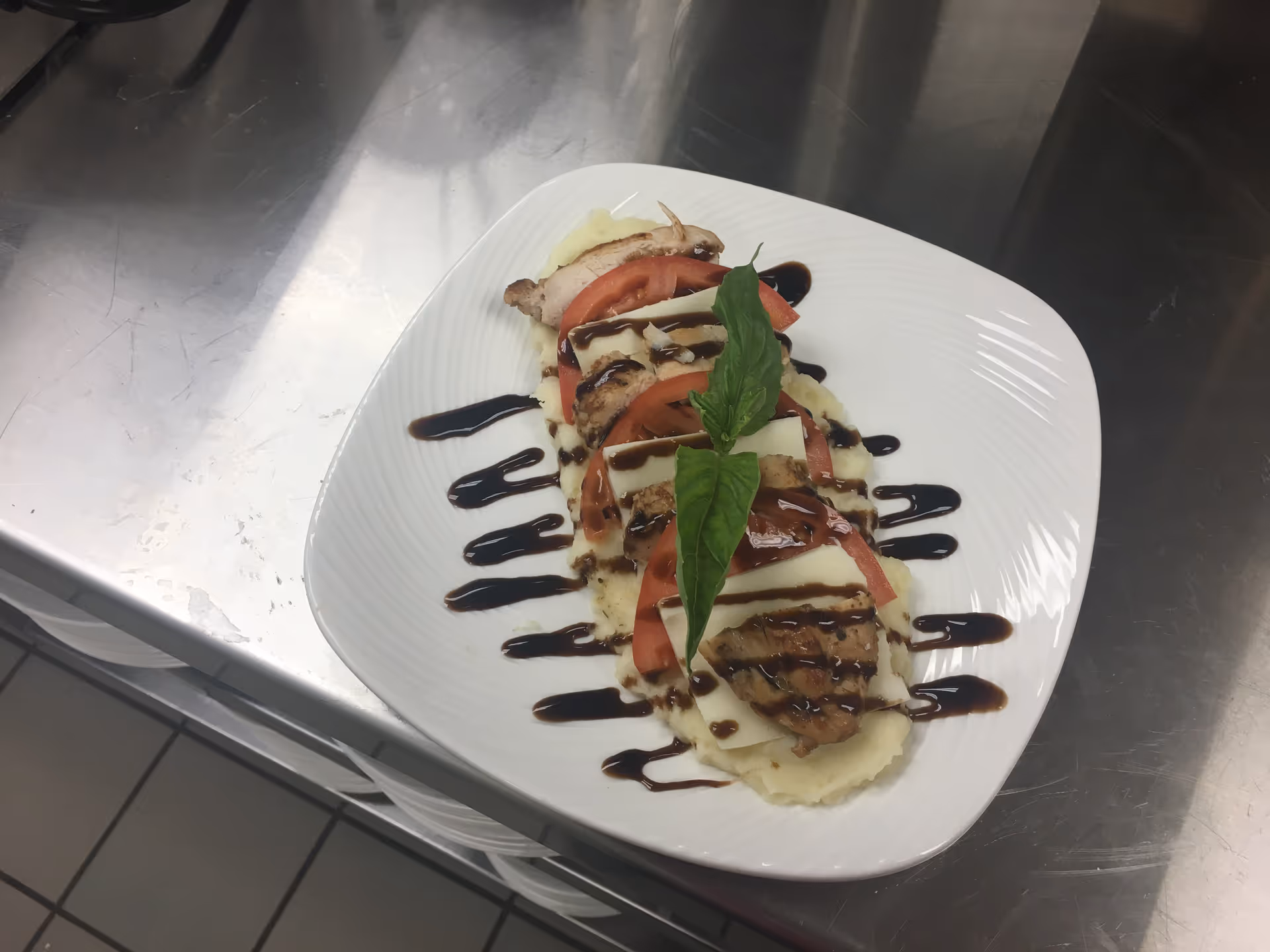 Plate of sliced meat with tomatoes, cheese, basil, and balsamic drizzle on a stainless counter