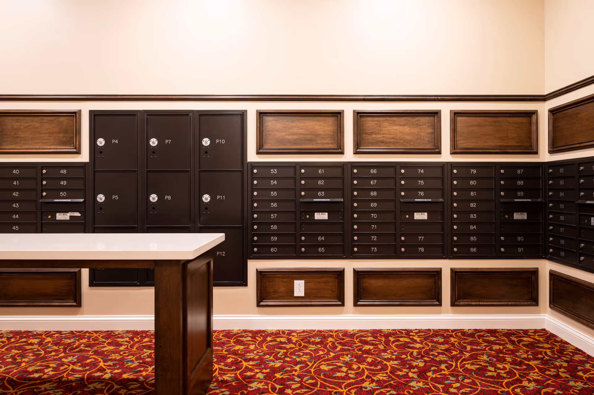 Interior view of a mailroom with multiple black mailboxes mounted on beige walls with wooden panel accents. A wooden table with a white countertop is positioned in front of the mailboxes. The floor is covered with a red patterned carpet.