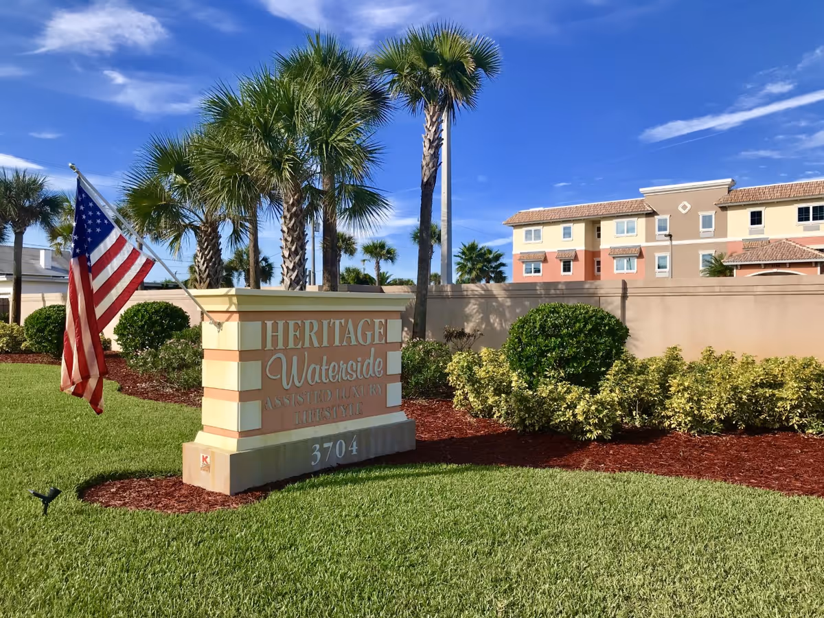 Outdoor view of Heritage Waterside Luxury Senior Living facility sign with an American flag mounted on it, surrounded by green grass, bushes, palm trees, and a clear blue sky. A multi-story building is visible in the background behind a beige wall.