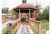 Wooden gazebo on a walkway in a grassy outdoor area beside a brick building.