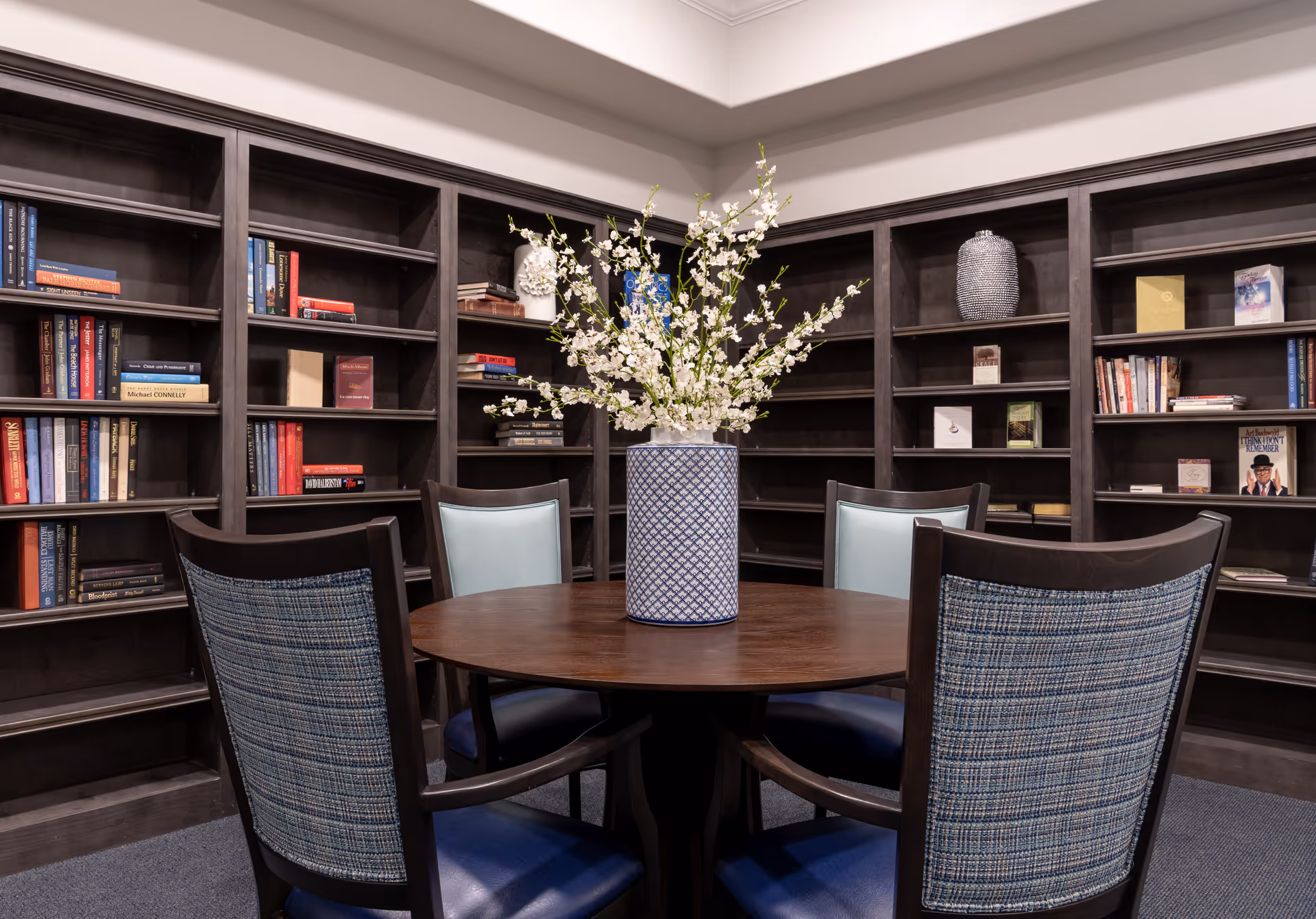 Round wooden table with a vase of white blossoms surrounded by upholstered chairs in a room lined with dark wood bookshelves.