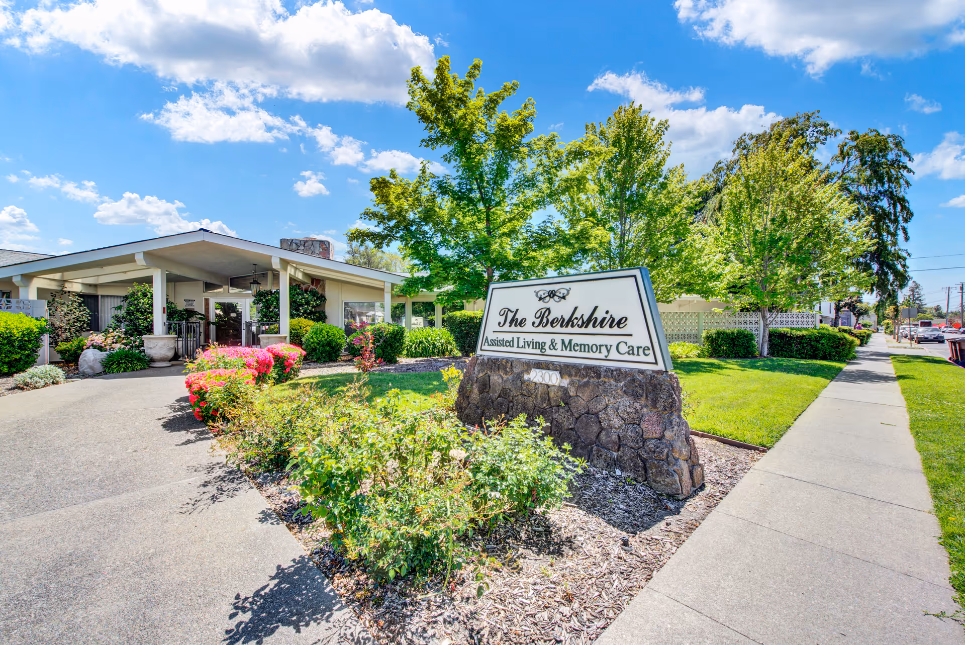 Exterior view of The Berkshire Assisted Living & Memory Care facility on a sunny day, showing a stone sign with the facility name, a pathway, green trees, bushes, and a building entrance.