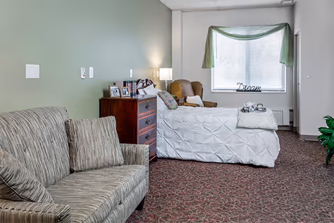 Interior of a senior living bedroom with bed, dresser, sofa, and window with green drape.