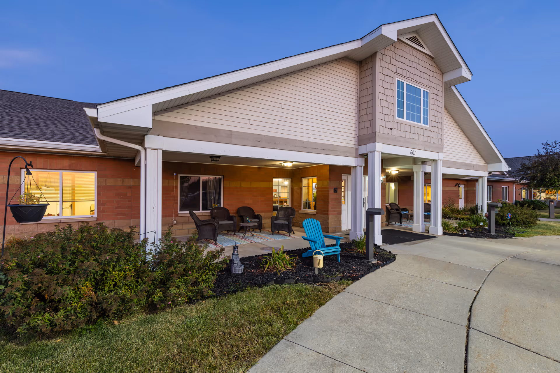 Exterior view of Courtyard Estates at Hawthorne Crossing building entrance during dusk, featuring a covered porch with outdoor seating including wicker chairs and a blue Adirondack chair, surrounded by landscaped bushes and a curved concrete walkway.
