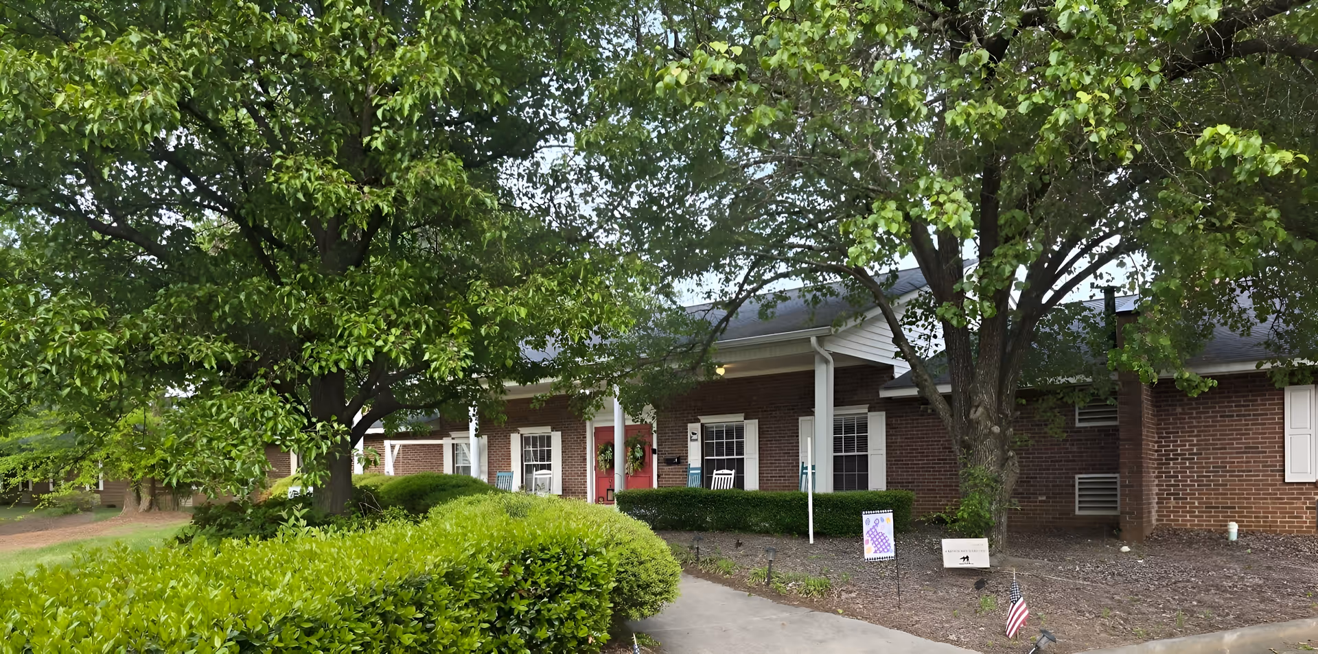 Brick assisted-living facility with a covered entrance, trees, and a paved walkway.