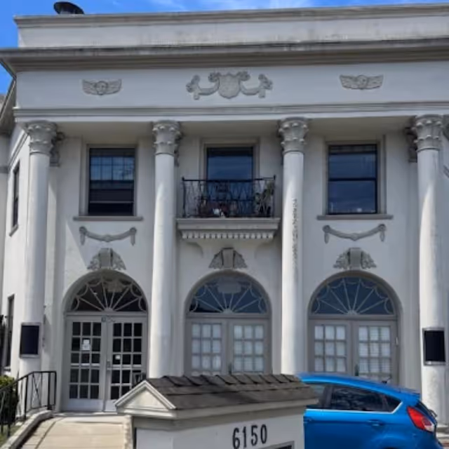 A white, classical-style building with tall columns, arched windows, and a blue car parked outside.