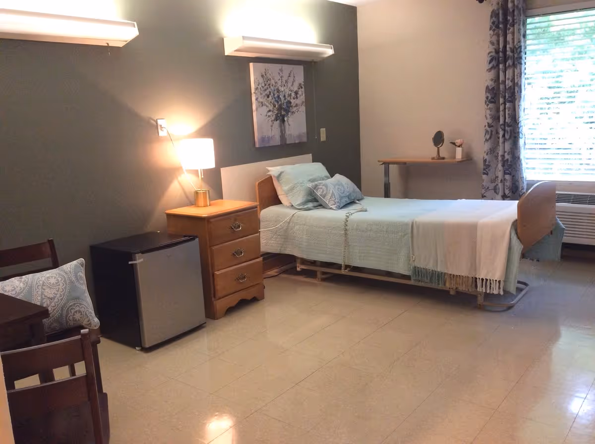 Senior living bedroom with a bed, nightstand, a dresser, and a window with curtains.